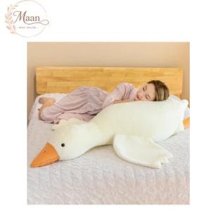 May include: A large, soft, cream-colored plush goose toy with an orange beak and small black eyes. The stuffed animal is elongated in shape, featuring a long neck and flat wings, designed to be used as a body pillow for resting or lounging.