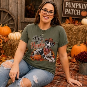 May include: An olive green short-sleeve t-shirt featuring a graphic of a black and white spotted dog wearing a brown cowboy hat and a red patterned bandana. The text "He's my Pumpkin" is printed above the dog, which is lying next to a pumpkin.