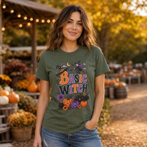 Può includere: Una t-shirt a maniche corte color verde oliva con una stampa grafica che riporta il testo "BASIC WITCH" in lettere arancioni e viola. Il design include un cappello da strega, una strega volante, pipistrelli, una luna crescente e varie zucche.