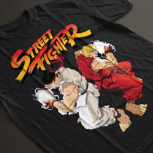 Può includere: Una t-shirt nera a maniche corte con una stampa grafica del logo Street Fighter in testo sfumato giallo e rosso sopra un'illustrazione di due artisti marziali in tenuta bianca e rossa che eseguono attacchi energetici luminosi.