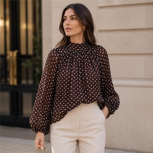 May include: A dark brown long-sleeved blouse featuring a white polka dot pattern, a high gathered neckline, and loose sleeves with elasticated cuffs. The garment is paired with light beige trousers, creating a contrast between the dark top and light bottoms.