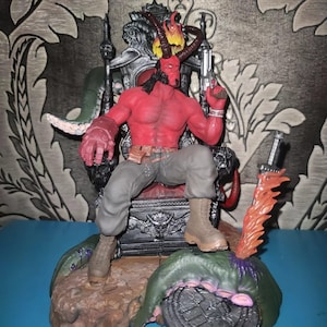 May include: A red-skinned Hellboy action figure with horns and a tail, sitting on a dark, ornate throne. The figure wears grey trousers and tan boots, holds a silver handgun, and features a flaming crown. A sword with orange flames stands nearby.