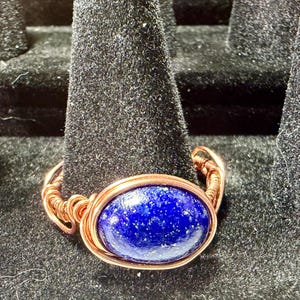May include: A ring featuring a smooth, oval-shaped deep blue lapis lazuli gemstone set within a polished copper wire-wrapped band. The metal wire is coiled around the stone and forms the shank of the ring, showcasing a metallic copper finish.
