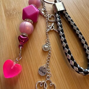 May include: A keychain featuring a braided black and silver strap attached to a metal ring. A strand of pink and fuchsia beads, including a star-shaped bead and a heart-shaped charm, hangs alongside silver chains with heart charms and a disc labeled LOVE.