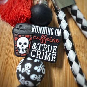 May include: A keychain featuring a black and white braided strap, a red tassel, and silicone beads. A central charm displays a coffee cup with a white skull and the text RUNNING ON caffeine & TRUE CRIME. Additional beads feature white skull patterns.