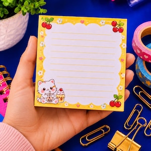 May include: A square notepad with a white lined centre and a yellow scalloped border decorated with small white flowers, red cherries, and a cartoon cat holding a boba tea drink. The notepad features a light yellow background with small hearts.