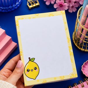 May include: A rectangular notepad with a white centre and a yellow border featuring a repeating lemon slice pattern. A cartoon lemon character with a smiling face, rosy cheeks, and a green leaf on top is printed in the bottom left corner of the page.