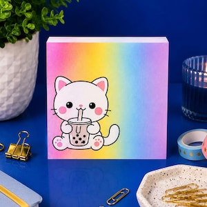 Peut inclure: Un bloc-notes carré avec un fond dégradé arc-en-ciel vibrant et une illustration centrale d'un chat blanc de dessin animé aux joues roses tenant une boisson bubble tea marron avec des perles de tapioca noires et une paille.