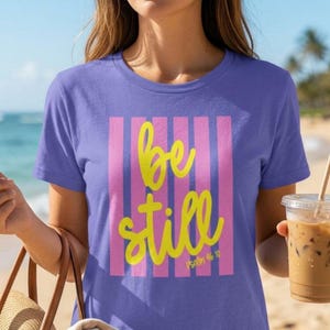Pode incluir: Uma camiseta lilás com um design de listras rosa verticais. As palavras "be still" estão escritas em amarelo, com "Psalm 46:10" abaixo. A camiseta é de manga curta.