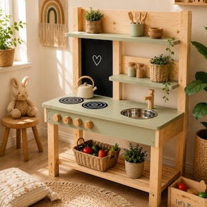 Op de afbeelding: Een houten speelkeuken met een saliegroen aanrechtblad, een roestvrijstalen spoelbak, een houten kraan en twee zwarte kookplaten. De unit bevat een zwart krijtbord achterpaneel met een witte harttekening, twee planken en een onderste opbergplank.