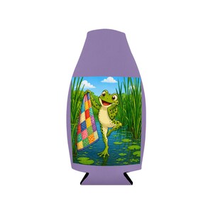 May include: A purple can cooler featuring a cartoon frog holding a colorful quilt in a pond setting. The frog is green with white spots and is smiling. The background includes tall green grass and a blue sky.