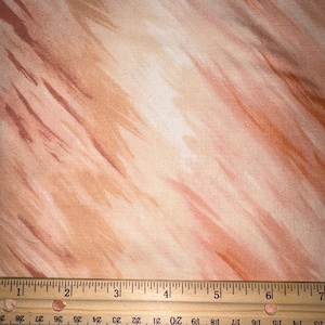 May include: A piece of fabric featuring a light peach and soft orange abstract brushstroke pattern. A wooden ruler is positioned along the bottom edge, showing a measurement scale in inches that spans approximately seven inches across the fabric.