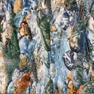 May include: A fabric featuring a realistic print of river stones in shades of blue, brown, and green with patches of grass, positioned above a wooden ruler showing a measurement of approximately 8.5 inches across the width of the material.