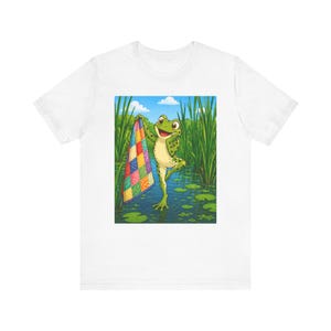 May include: White t-shirt featuring a cartoon frog holding a colorful patchwork quilt in a pond. The frog is green with a smiling face. The background shows tall green reeds, lily pads, and a blue sky with clouds. The quilt has squares of various colors.