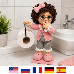 Puede incluir: Una muñeca de ganchillo con pelo rizado castaño oscuro, un lazo rosa y gafas redondas. La muñeca lleva una rebeca rosa con puños de volantes blancos sobre un mono gris y zapatillas rosas. Sostiene una varilla de madera con papel higiénico.