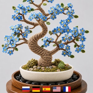 Op de afbeelding: Een handgehaakte bonsai boom met blauwe bloemen en groene bladeren. De boom heeft een gedraaide bruine stam en staat in een witte keramische pot met zwarte stenen. De pot staat op een houten dienblad.