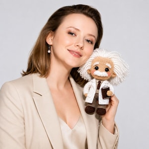 May include: A crocheted doll of Albert Einstein, wearing a white lab coat, brown trousers, and holding a book. The doll has white hair and a moustache. The doll is being held by a person wearing a beige blazer.