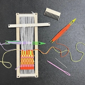 Può includere: Un telaio rettangolare in legno con fili di ordito bianchi, un bastoncino per l'apertura del passo e un tessuto parzialmente intrecciato in arancione, giallo e rosa. Include un pettine in legno, una navetta verde in plastica con filo viola, un ago arancione e uno rosa.