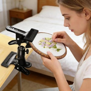 May include: A black adjustable embroidery hoop stand with a heavy-duty clamp mechanism for table mounting. The stand features a ball-and-socket joint for positioning and holds a round wooden embroidery hoop containing a floral needlepoint design.