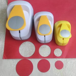 May include: Three white and yellow paper punches of varying sizes arranged in a row. Each punch features a gray circular button on top. Below the punches, three red paper circles of corresponding sizes are displayed on a red sheet of paper.