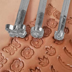 Könnte beinhalten: Drei silberne Metallstempelwerkzeuge für Leder mit strukturierten Griffen, jedes mit einem eigenen Motiv an der Spitze, darunter ein Schmetterling, eine mehrblättrige Blume und eine kleine Blütenform, eingedrückt in eine Lederfläche.