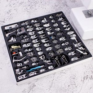 May include: A comprehensive set of silver-toned metal sewing machine presser feet organized in a black plastic tray. The collection includes various specialized attachments for different stitching tasks, featuring metallic finishes and clear plastic components.