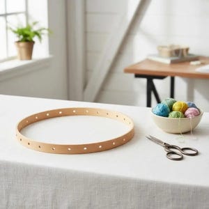 May include: A circular wooden hoop with evenly spaced holes along its perimeter, accompanied by a small cream-coloured bowl filled with various colourful yarn balls and a pair of metal scissors resting on a white fabric surface.