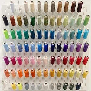 May include: A white pegboard display rack holding a large collection of sewing thread spools in a wide spectrum of colors. The spools are arranged in neat rows and columns, showcasing shades of white, brown, green, blue, purple, pink, red, and yellow.