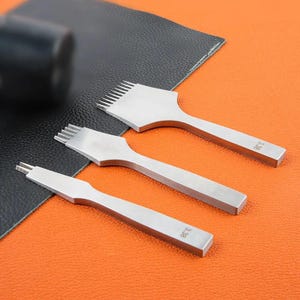 May include: Three silver-colored metal leather stitching chisels with varying prong counts, each featuring the number 3.38 engraved on the handle. These tools are arranged diagonally on a black textured surface against an orange background.