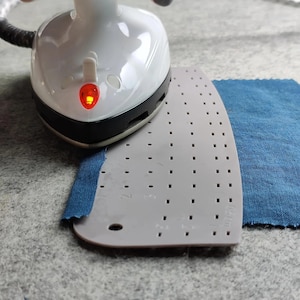 Puede incluir: Una plancha de mano blanca y negra con una luz indicadora roja encendida, colocada sobre una regla de planchado de plástico gris. La regla presenta una cuadrícula de pequeños orificios y marcas impresas, incluyendo el texto 0.5 cm, sobre tela azul.
