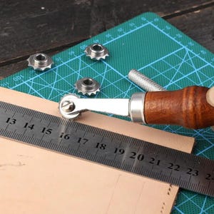 May include: A leather stitching groover tool with a wooden handle and a metal wheel, positioned on a piece of light tan leather next to a metal ruler showing measurements in centimetres. Three additional interchangeable metal wheels are nearby.