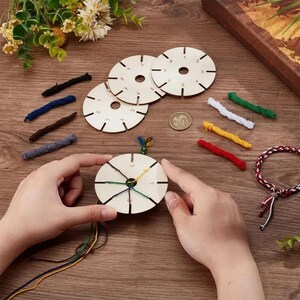 May include: Three circular wooden kumihimo braiding discs with numbered slots, accompanied by several short lengths of coloured embroidery thread, a small metallic coin, and a partially completed braided bracelet on a wooden surface.