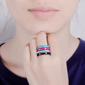 May include: A stack of four thin metal rings featuring smooth enamel bands in bright turquoise, hot pink, light blue, and black. Each ring is accented with a single small, clear, round-cut stone set into the centre of the coloured enamel surface.