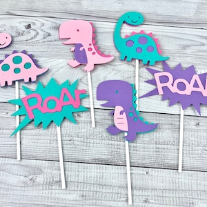 K&ouml;nnte beinhalten: Sechs bunte Dinosaurier-Kuchentopper, darunter zwei rosa Dinosaurier, zwei lila Dinosaurier, ein t&uuml;rkisfarbener Dinosaurier und ein t&uuml;rkisfarbenes "Roar"-Schild. Die Dinosaurier haben rosa, lila und t&uuml;rkisfarbene Akzente.