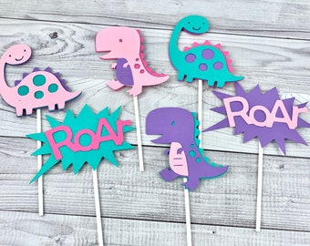 May include: Six colorful dinosaur cake toppers, including two pink dinosaurs, two purple dinosaurs, one teal dinosaur, and one teal "Roar" sign. The dinosaurs have pink, purple, and teal accents.