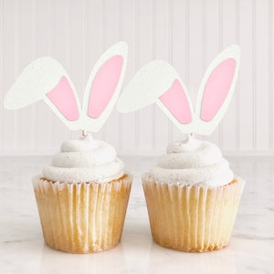 Könnte beinhalten: Zwei weiße Cupcakes mit Vanille-Frosting, verziert mit rosa und weißen Cupcake-Toppern in Form von Hasenohren.