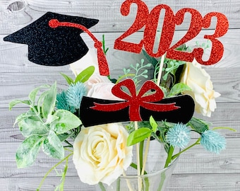 Puede incluir: Centro de mesa de graduación con un gorro de graduación negro, un diploma rojo con un lazo rojo y un 2023 de purpurina roja. Las decoraciones se colocan en un jarrón de vidrio transparente con flores blancas y azules.