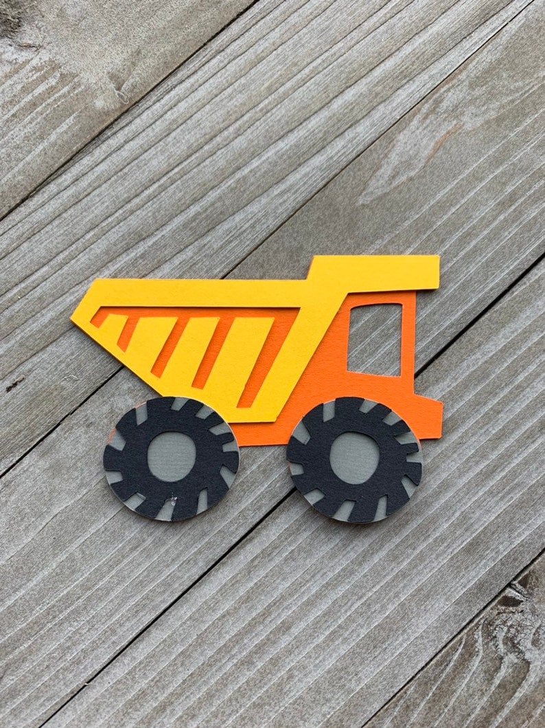 May include: A yellow and orange paper cut-out of a dump truck with black tires. The truck is on a wood background.
