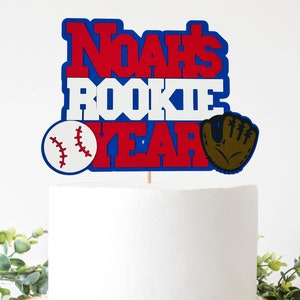 May include: A red, white, and blue cake topper with the text "Noah's Rookie Year" and a baseball and baseball glove.
