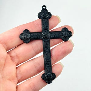 Könnte beinhalten: Ein schwarzer, verzierter Kreuzanhänger mit einem detaillierten Design. Das Kreuz ist aus Kunststoff und hat eine glatte Oberfläche.