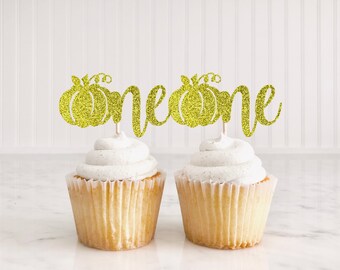 Peut inclure: Deux cupcakes avec un glaçage blanc surmontés de décorations de cupcakes en forme de citrouille dorées à paillettes qui disent "one".