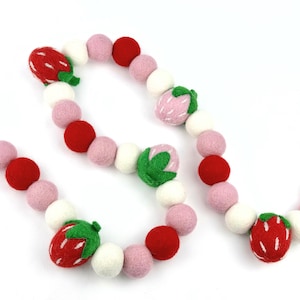 Puede incluir: Una guirnalda de bolas de fieltro en rosa, rojo y blanco con detalles de fresa. La guirnalda está hecha de bolas de fieltro y presenta un diseño de fresa.