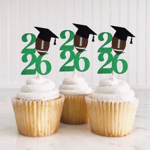 Può includere: Tre cupcakes sormontati da decorazioni glitterate verdi "2026", ognuna con un pallone da football e un cappello di laurea. I cupcakes hanno una glassa bianca e sono in pirottini di carta marrone chiaro. Sfondo bianco strutturato.