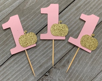 Peut inclure: Trois décorations de cupcake roses avec le chiffre "1" et une citrouille dorée pailletée sur chacune.