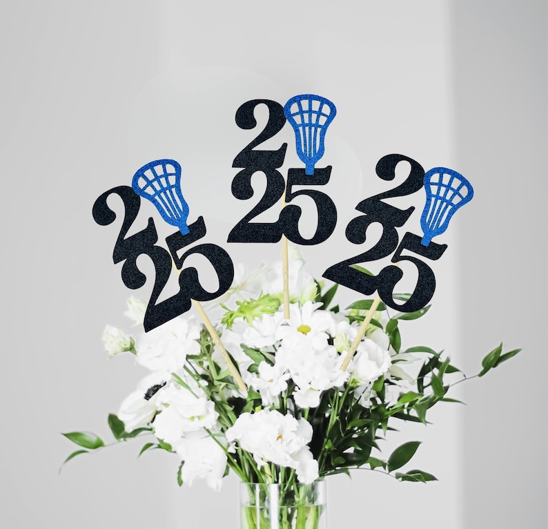 May include: Black glittery number 25 centerpieces with blue lacrosse stick accents on wooden sticks. The centerpieces are arranged in a vase of white flowers and greenery, perfect for a graduation celebration.
