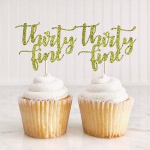 May include: Two cupcakes with white frosting topped with gold glitter cupcake toppers that say "thirty fine".