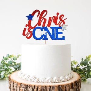 Puede incluir: Topper de pastel de purpurina roja y azul con el texto "Chris is ONE" y estrellas plateadas. El topper está en un pastel blanco con glaseado blanco.