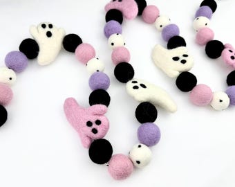 May include: A Halloween-themed garland featuring felt ghost and pink monster shapes, interspersed with black, white, and lavender felt balls. The garland is strung together with a thin black and white string.
