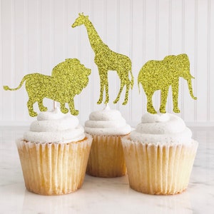 Könnte beinhalten: Drei goldene Glitzer-Tier-Cupcake-Topper, ein Löwe, eine Giraffe und ein Elefant, auf weißen, mit Zuckerguss überzogenen Cupcakes.