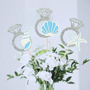 May include: Three decorative toppers on wooden sticks featuring silver glitter diamond ring cutouts. Each ring is accented with a different sea-themed shape: a white and blue spiral shell, a light blue scallop shell, and a cream-colored starfish.